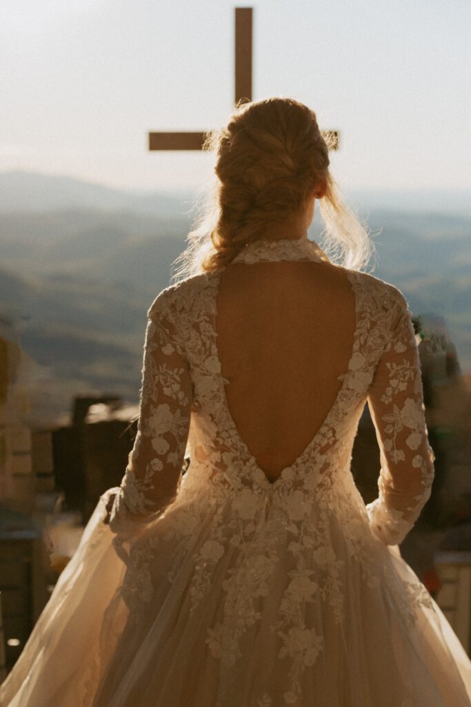 Woman in wedding dress walking towards cross and mountains.