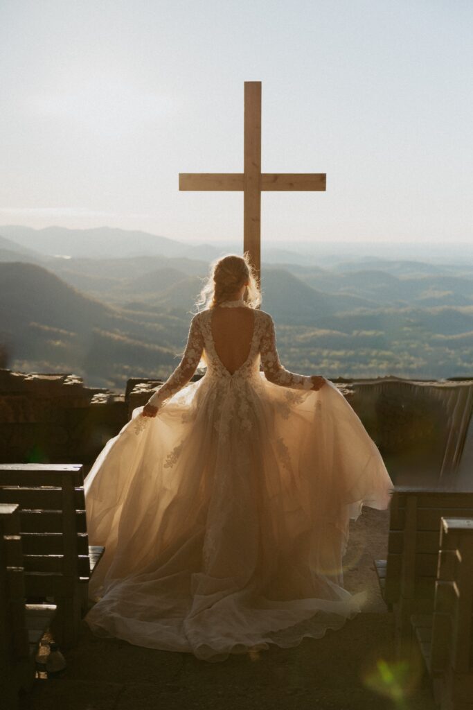 Woman in wedding dress walking down steps toward a cross with mountains behind it. 