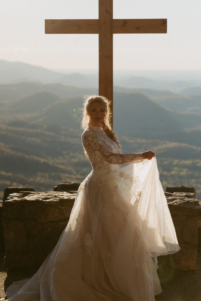 Woman in wedding dress standing in front of cross and mountains.