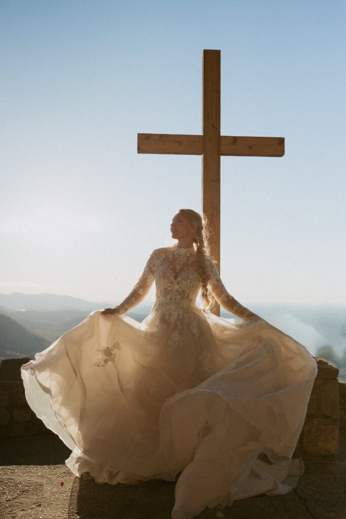 Woman in wedding dress spinning in front of cross with mountains in the background. 