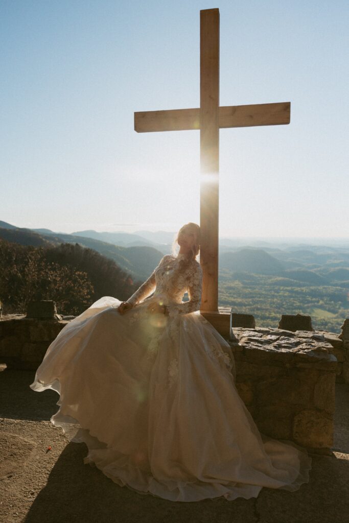 Woman in wedding dress sitting in front of cross with mountains in the background. 