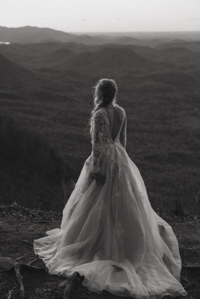Woman in wedding dress standing on rock looking at mountains.