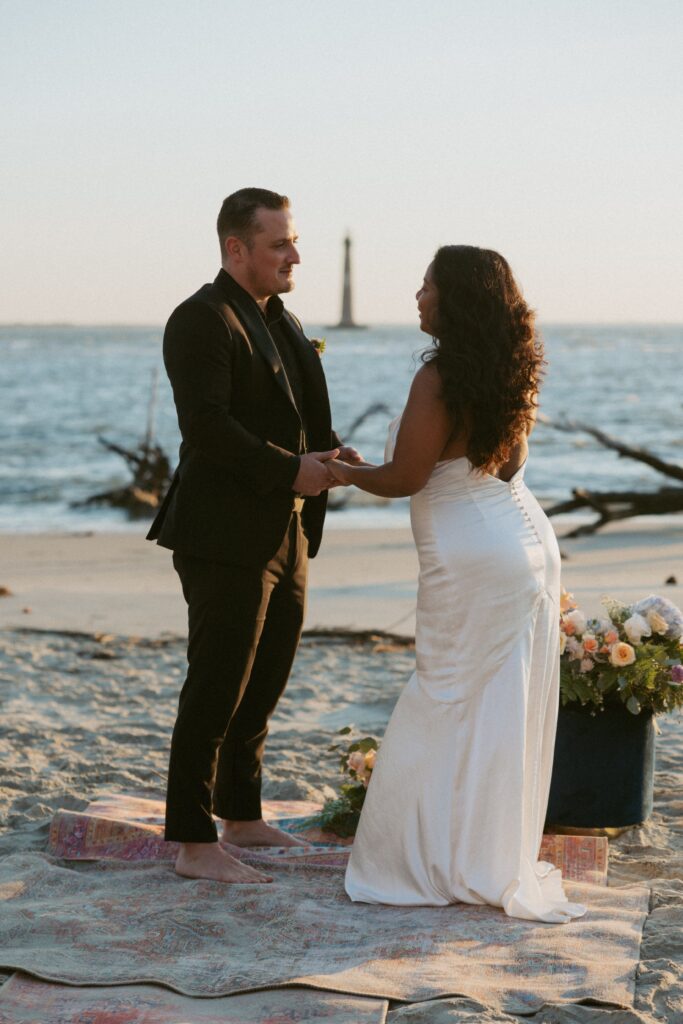 Couple in wedding attire standing on beach and holding hands with ocean and lighthouse behind them.