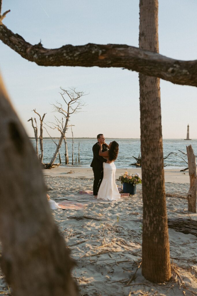 Man and woman in wedding attire dancing on a rug on the beach with trees in the foreground.