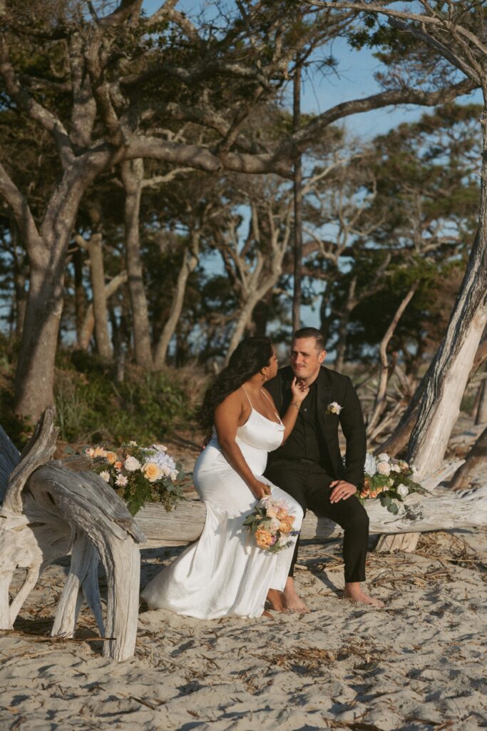 Man and woman in wedding attire sitting on driftwood on the beach with flowers around them.