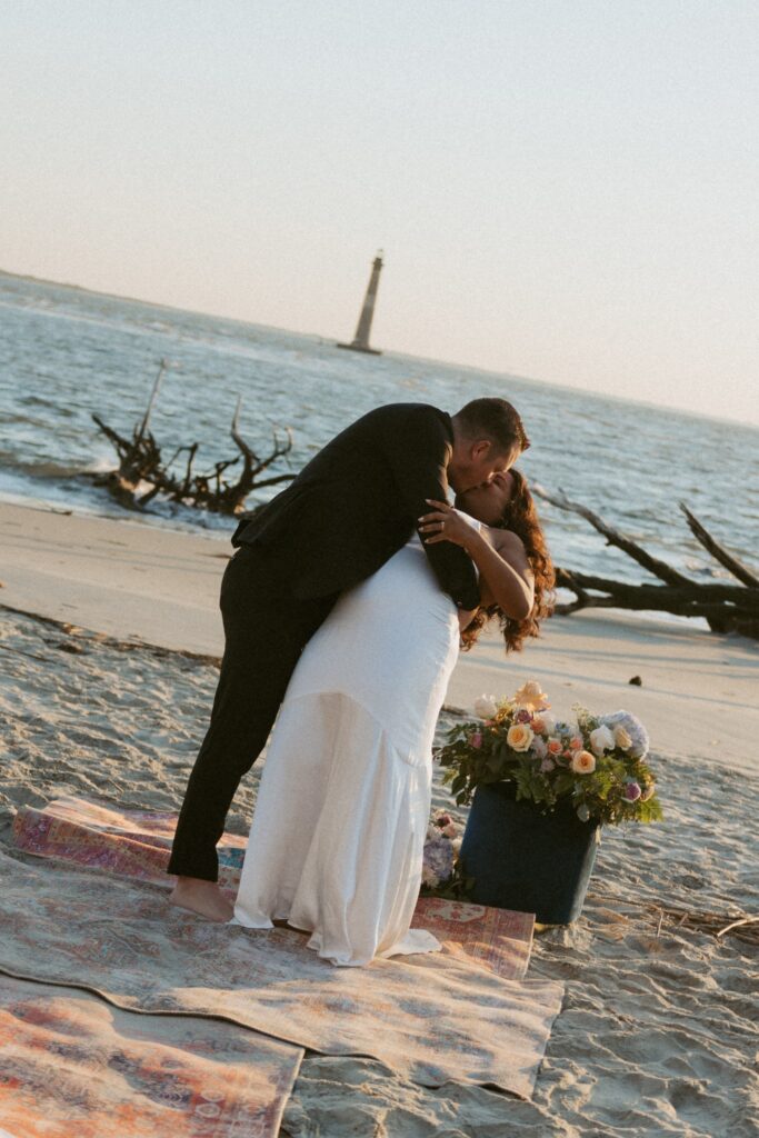 Man and woman in wedding attire kissing while standing on a rug on the beach. 