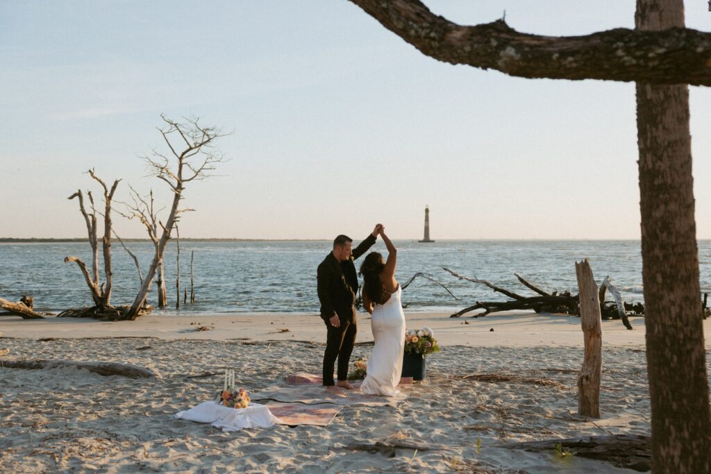 Man in a suit spinning woman in wedding dress while standing on a rug on the beach with ocean and lighthouse behind them and trees in the foreground.