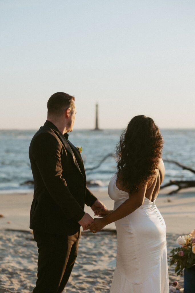 Man and woman in wedding attire looking at ocean and lighthouse while holding hands.