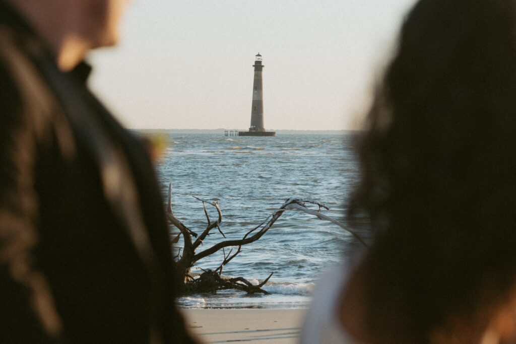 Man and woman in foreground looking at lighthouse in the ocean. 