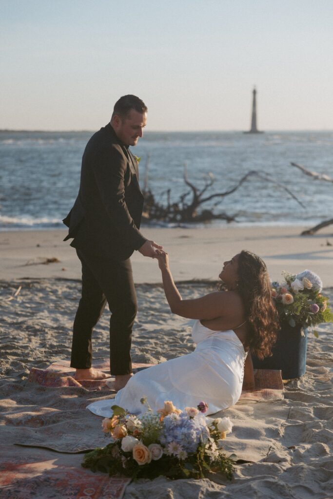 Woman in wedding dress sitting on rug on the beach while man is standing in suit and helping her up. 