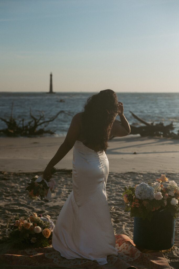 Woman in wedding dress standing on rug and looking at ocean while holding a bouquet of flowers.