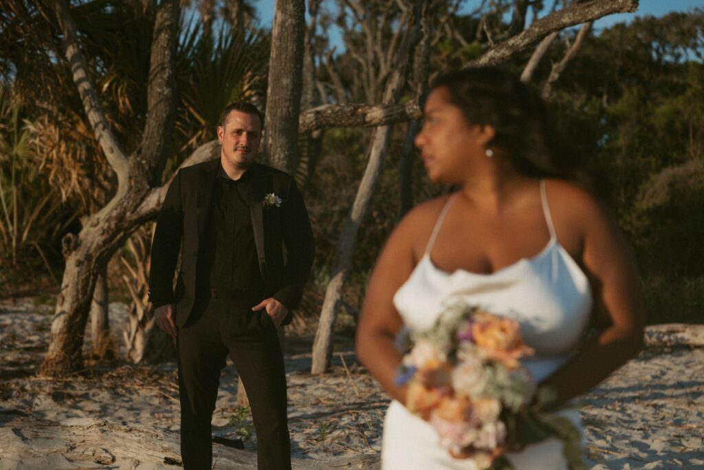 Woman in wedding dress holding a bouquet in the foreground with man in the background in suit looking at her.