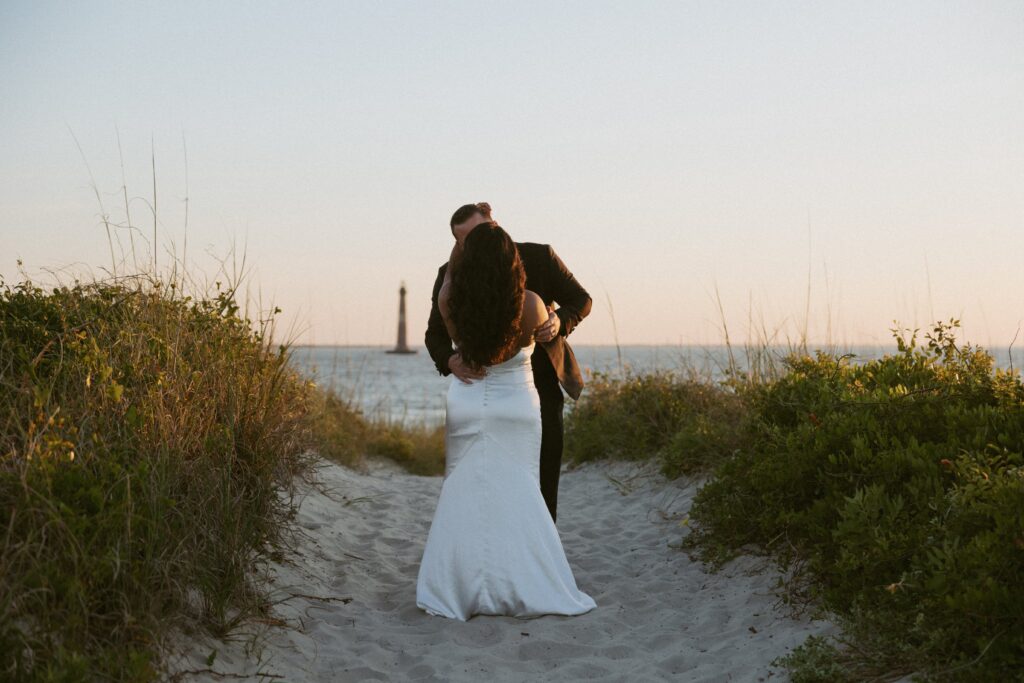 Man and woman in wedding attire hugging and kissing on the beach with ocean and lighthouse behind them.