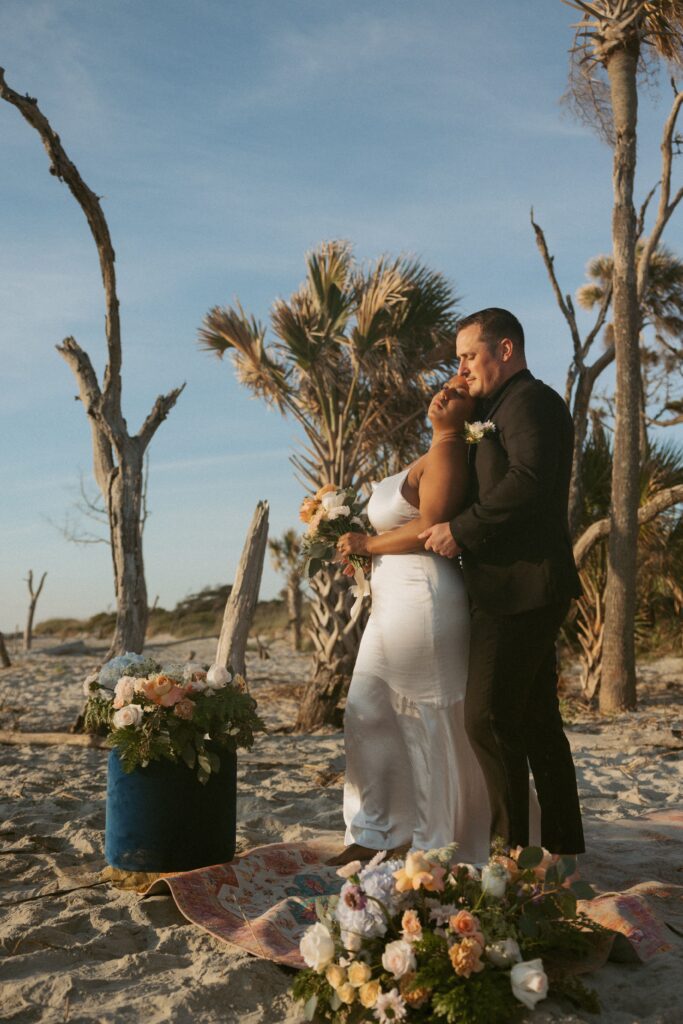 Man and woman in wedding attire cuddling while standing on a rug on the beach.