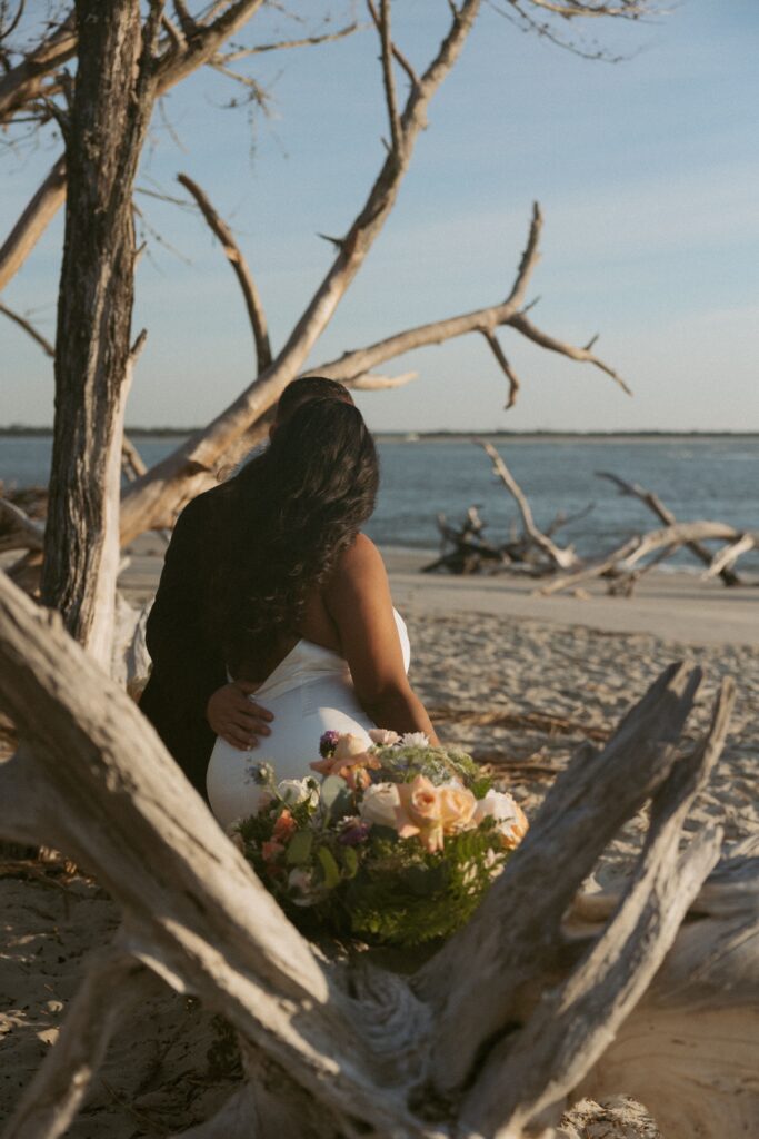 Man and woman in wedding attire sitting on driftwood on the beach with colorful florals beside them.