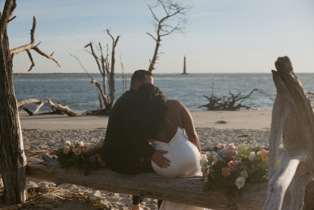 Man and woman sitting on a piece of driftwood looking at the ocean with colorful flowers beside them.