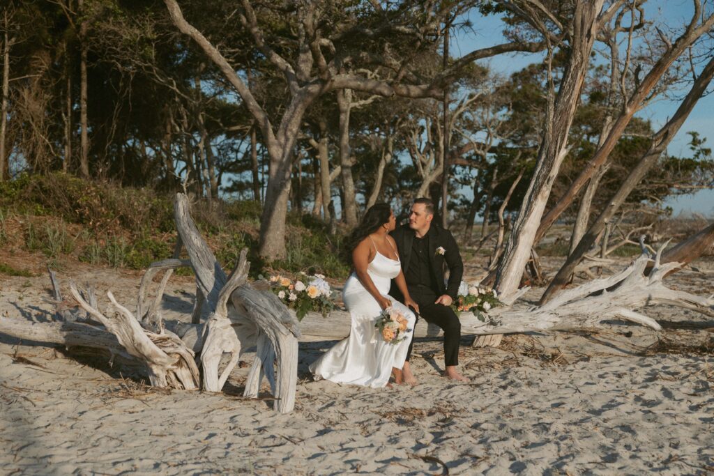 Man and woman in wedding attire sitting on driftwood on the beach with colorful flowers around them.