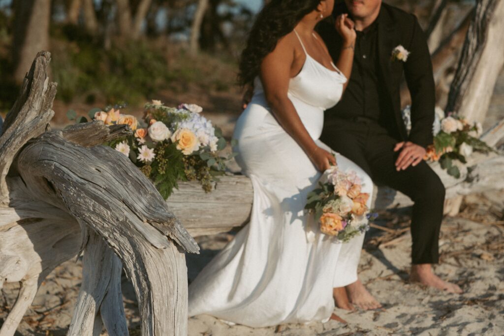 Man and woman in wedding attire sitting on driftwood in with colorful flowers around them.