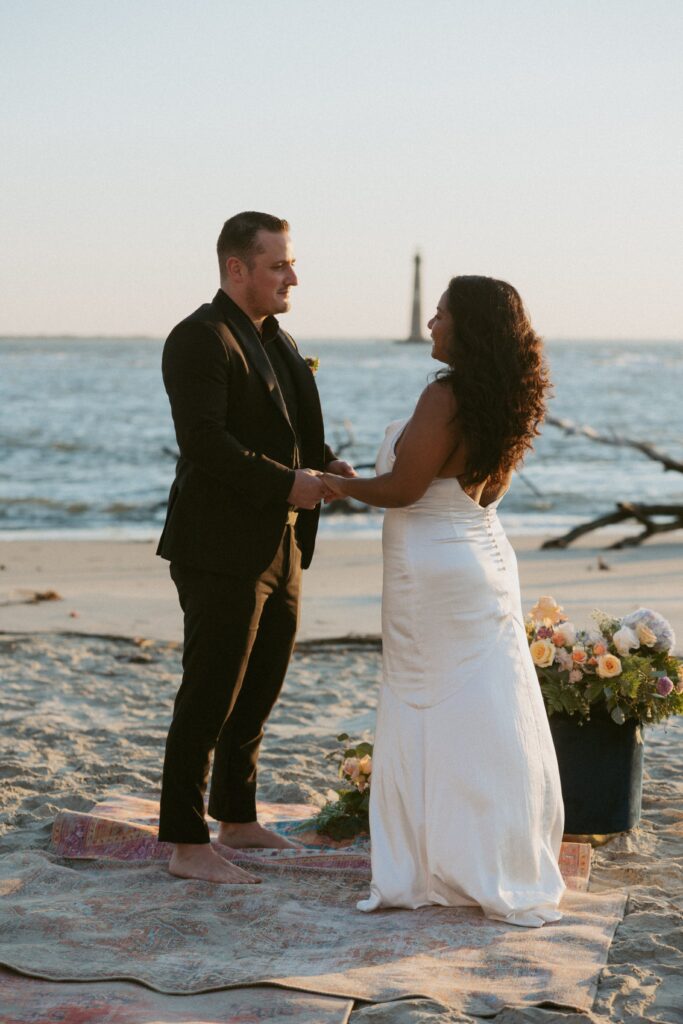 Man and woman in wedding attire looking at each other and holding hands while standing on a rug on the beach. 