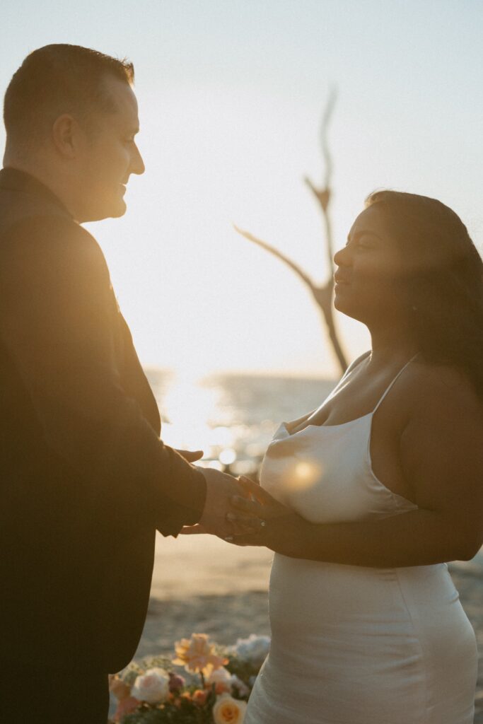 Man and woman in wedding attire holding hands on beach with sun shining between them.