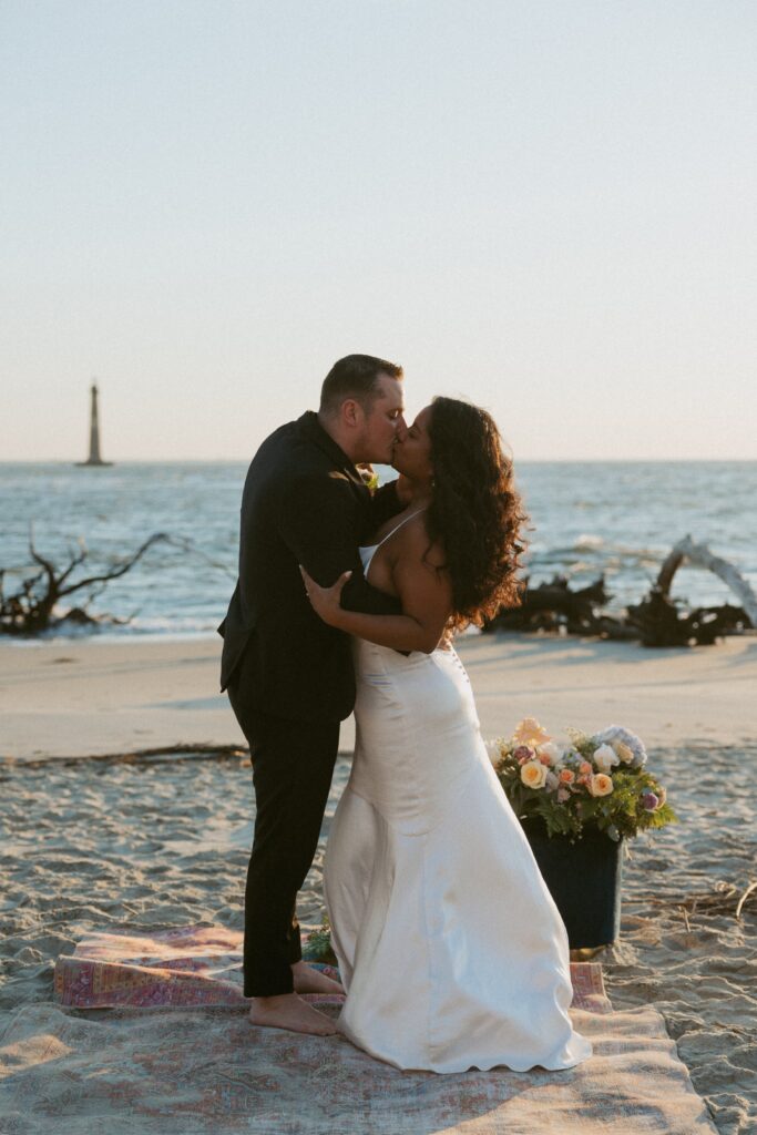 Man and woman standing on rug on beach in wedding attire and kissing. 