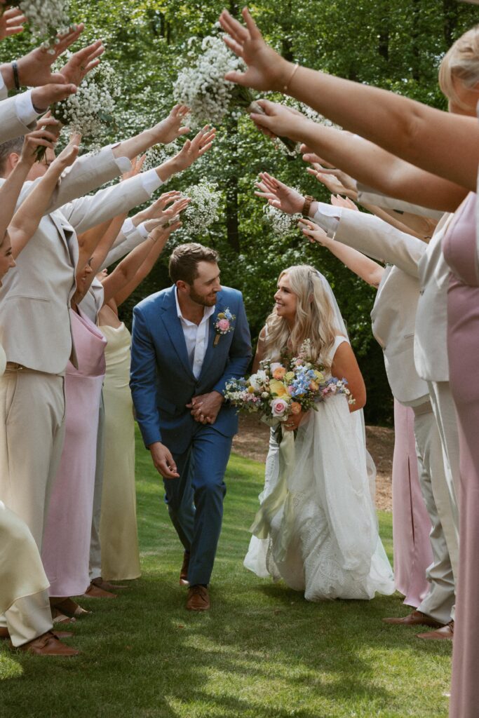 Man and woman in wedding attire walking through tunnel of bridal parties hands and flowers.