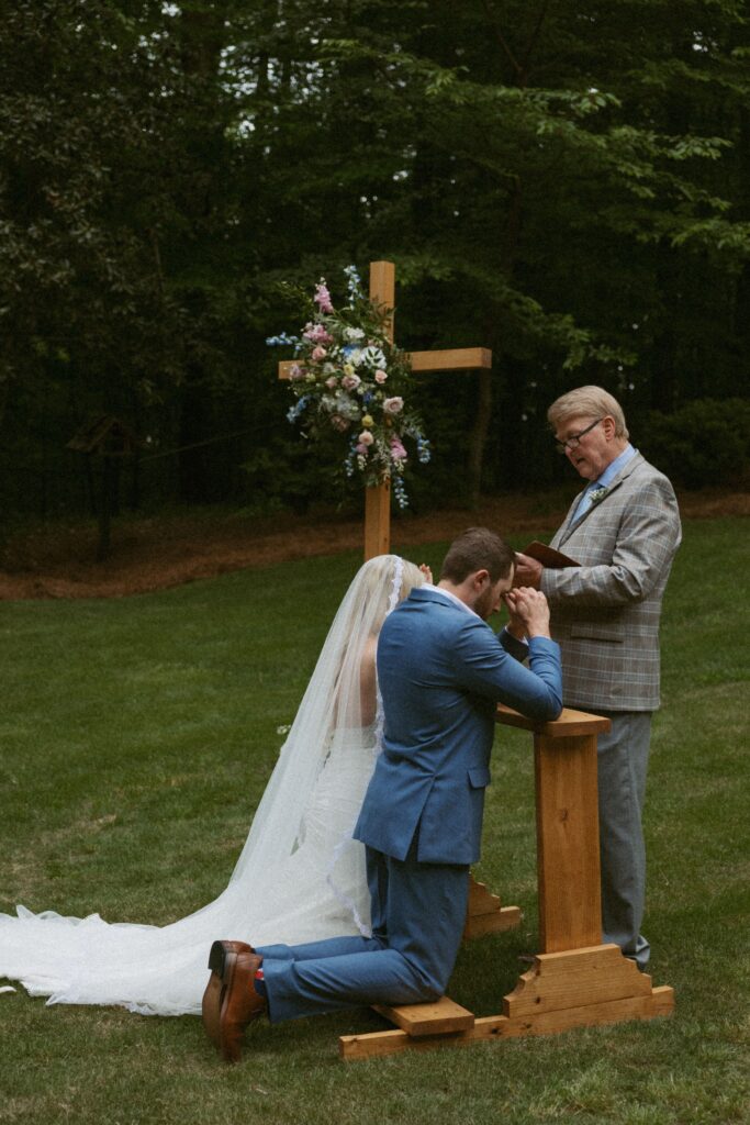 Man and woman kneeling at an alter in the backyard with an officiant reading out of the bible. 