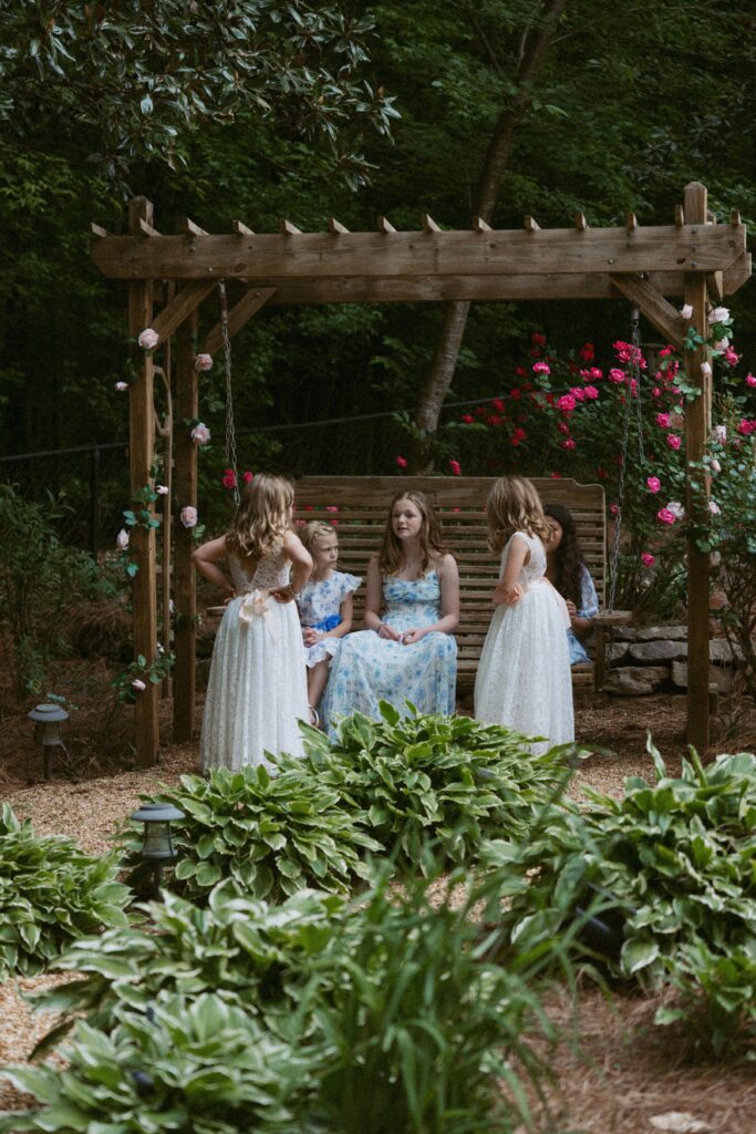 Little girls in dresses standing and sitting on a swing in a backyard garden. 