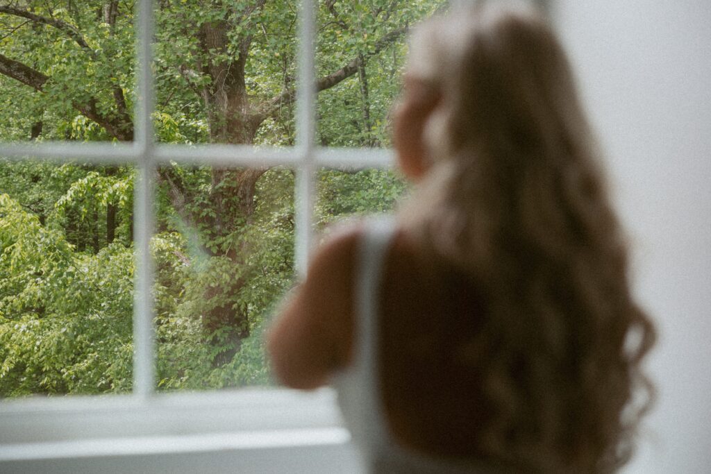 Woman in wedding dress looking out window at trees in the yard.