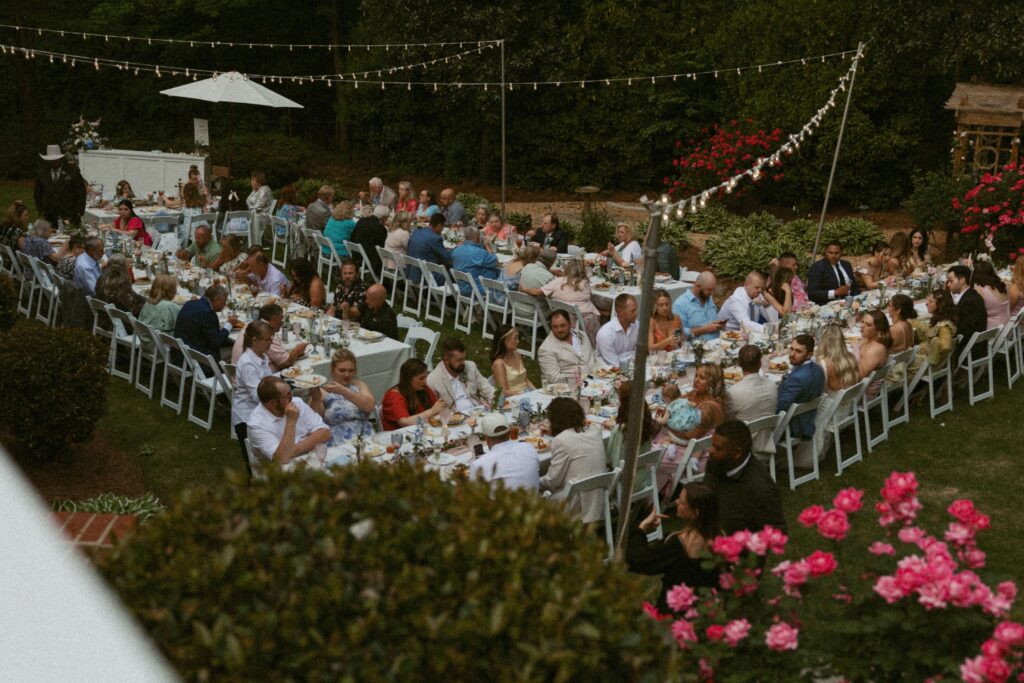 Group of people sitting at large tables and eating with lights and flowers all around them.