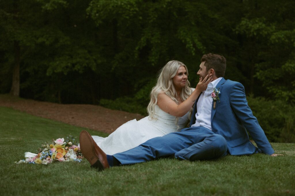 Man and woman in wedding attire sitting on the grass and smiling at each other.