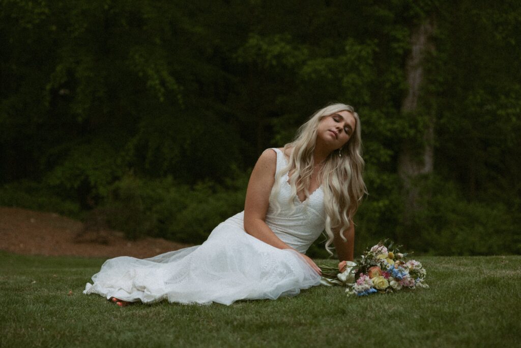 Woman in wedding dress sitting in grass with a flower bouquet sitting next to her.