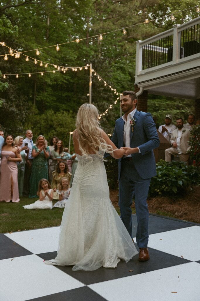 Man and woman in wedding attire dancing on a black and white checkered dance floor with a crowd of people watching them.