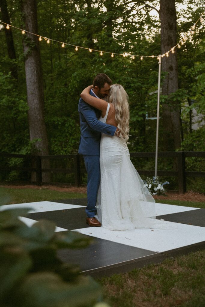 Man and woman in wedding attire slow dancing on a white and black checkered dance floor with lights above them and greenery in the foreground.