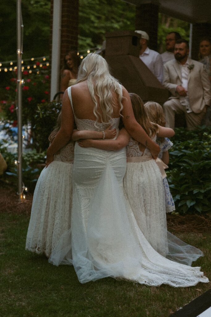 Woman in wedding dress from behind while she is hugging flower girls.