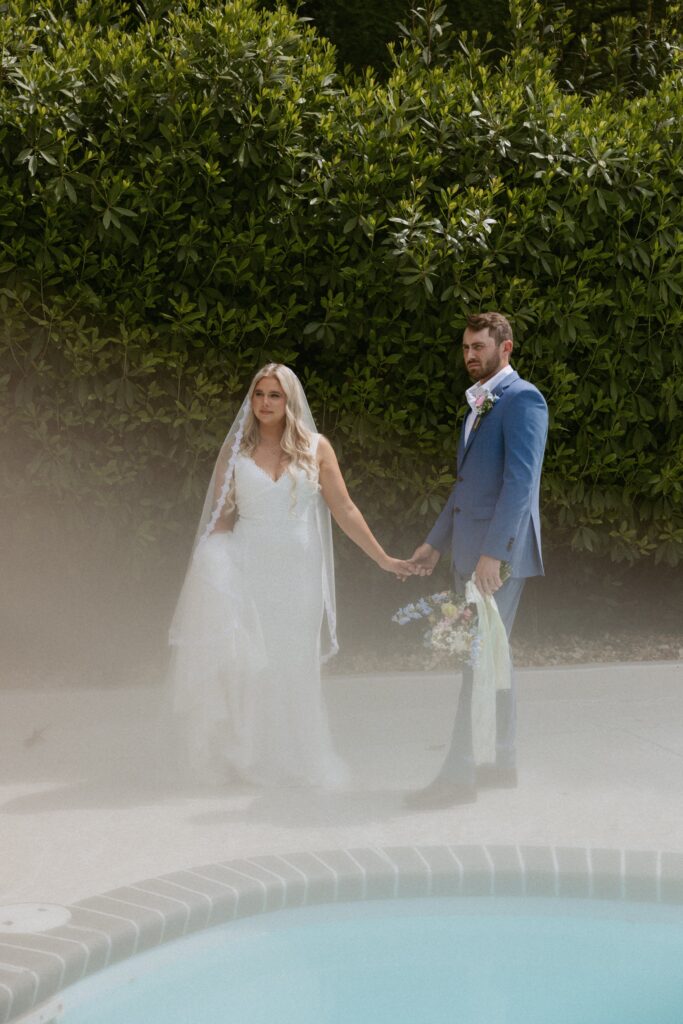 Man and woman in wedding attire holding hands standing beside a pool.