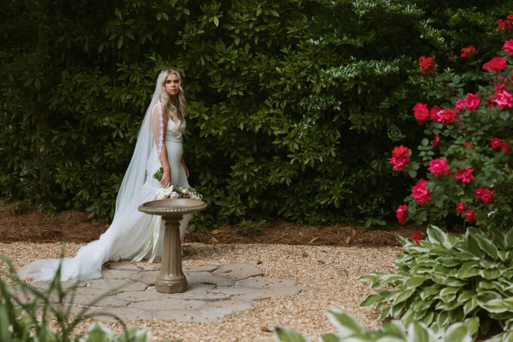 Woman in wedding dress with veil and flowers walking around water fountain in garden.