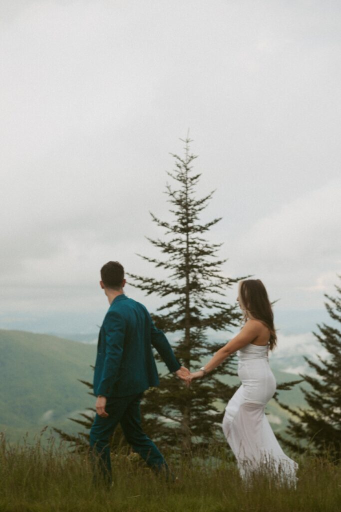 Man and woman holding hands and walking through grass with mountains and trees behind them.