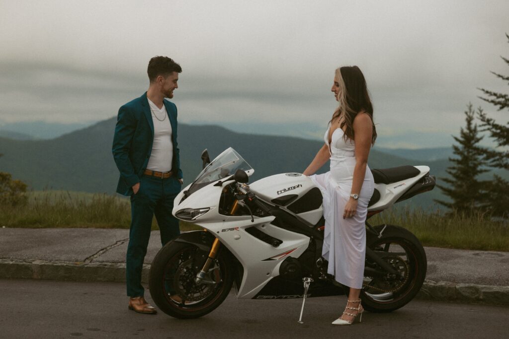 Woman sitting on motorcycle and man standing beside motorcycle with mountains in the background.