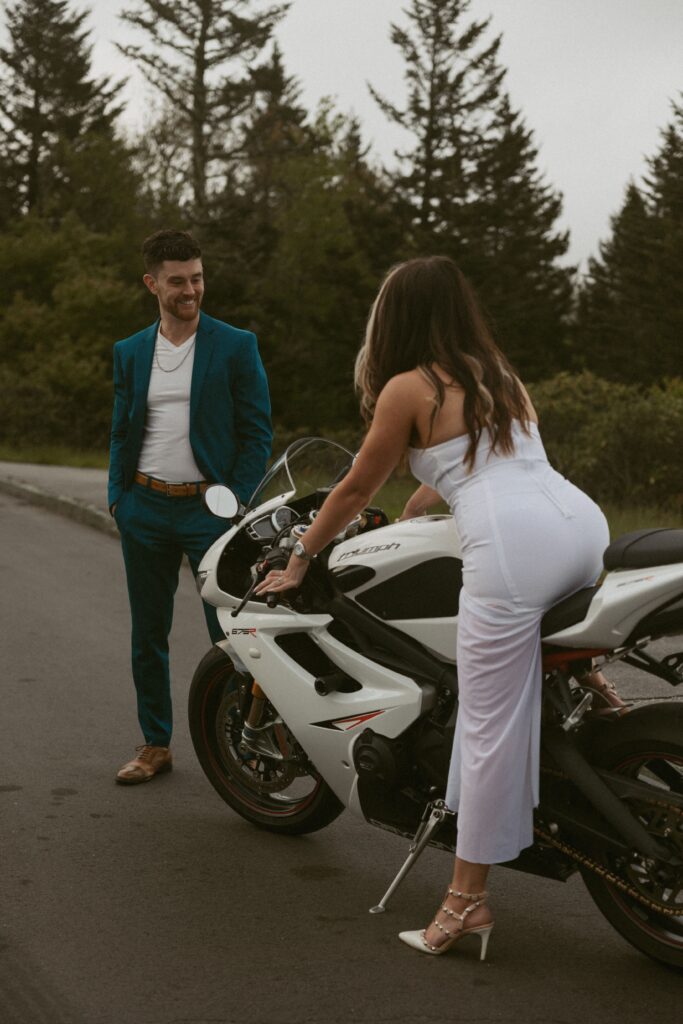 Woman sitting on motorcycle while man looks and smiles at her.