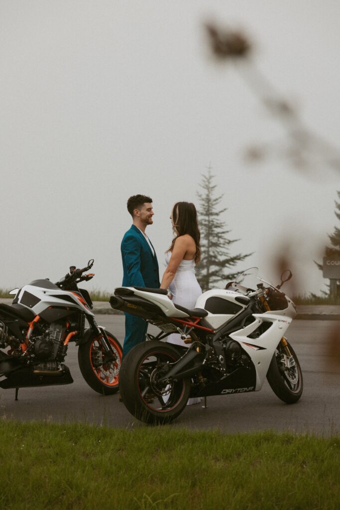 Man and woman smiling and looking at each other while they are standing between motorcycles.