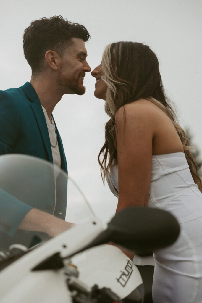 Man and woman leaning over a motorcycle for a kiss but smiling.