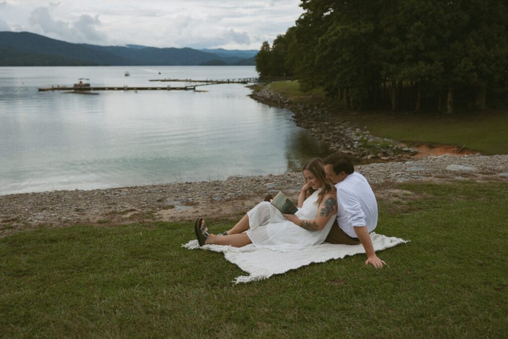 Man and woman sitting on a blanket in the grass while reading a book. 