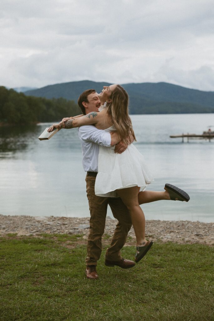 Man picking a woman up and spinning her while she is holding a book and laughing in front of a lake. 