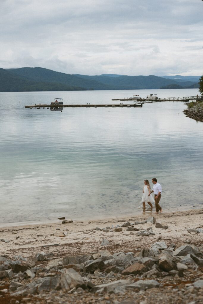 Man and woman walking down the beach at a lake with the mountains in the background.