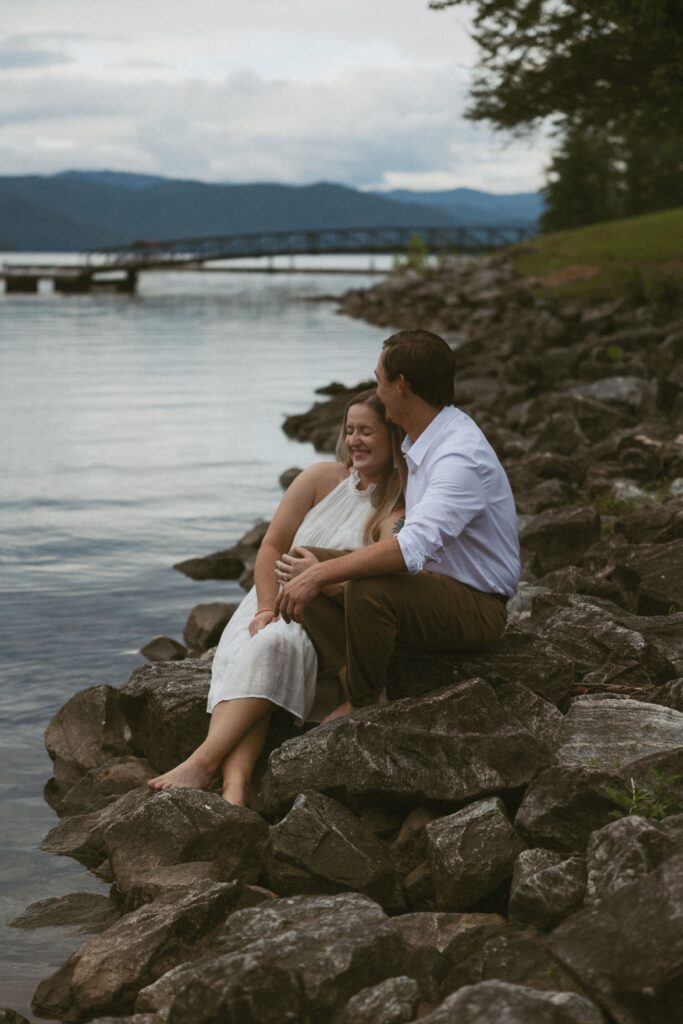 Woman smiling and sitting in between mans legs on the rocks beside a lake. 