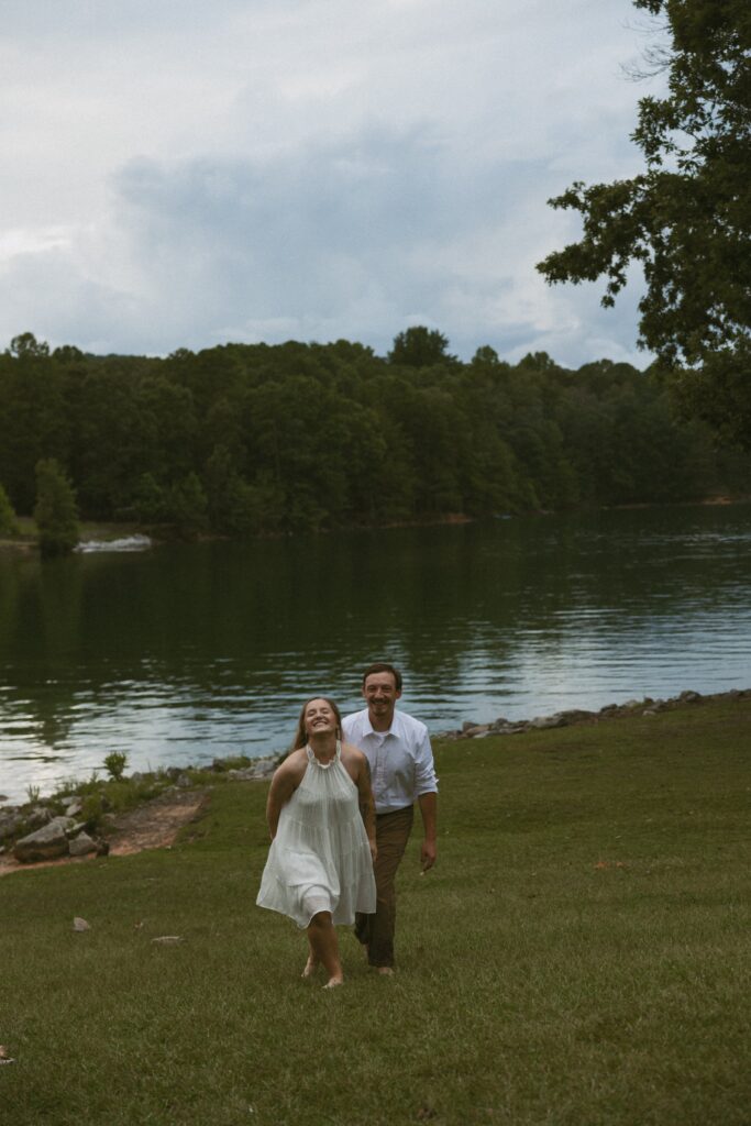 Man and woman walking through the grass in front of a lake and laughing. 