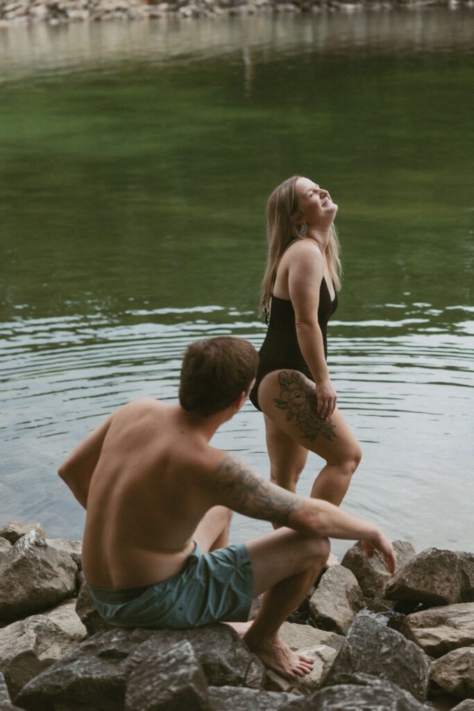 Woman standing in the water laughing while a man is sitting on the rocks watching her. 