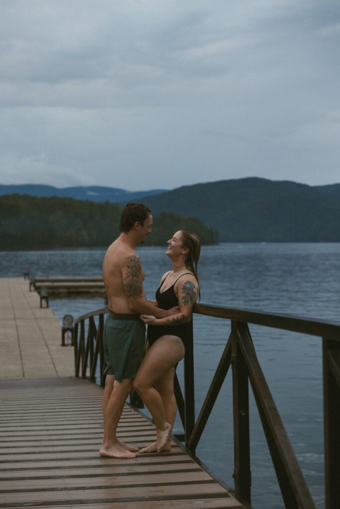 Man and woman leaning against the rail on a dock with the lake and mountains behind them.