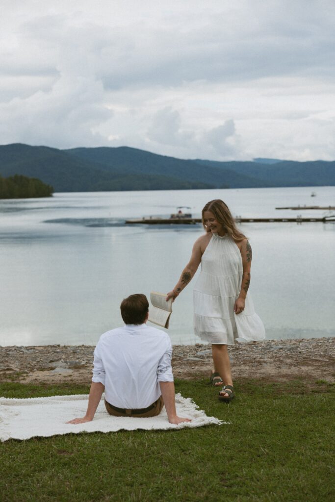 Woman holding a book while walking around a man sitting on the grass with mountains behind them. 